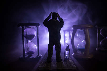 Time concept. Silhouette of a man standing between hourglasses with smoke and lights on a dark...