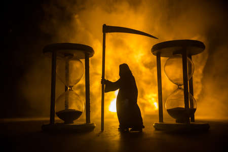 Time concept. Scary view of grim reaper silhouette standing between hourglasses with smoke and lights on a dark background. Surreal decorated pictureの写真素材