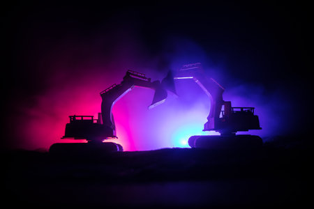 Construction site on a city street. A yellow digger excavator parked during the night on a construction site. Industrial concept table decoration on dark foggy toned background. Selective focusの写真素材