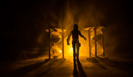 Time concept. Silhouette of a man standing between hourglasses with smoke and lights on a dark background. Surreal decorated pictureの写真素材