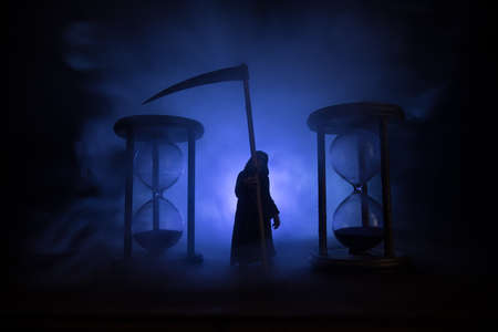 time concept. Scary view of grim reaper silhouette standing between hourglasses with smoke and...