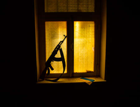 Conceptual photo of war between Russia and Ukraine. Ukrainian flag, helmet and gun on windowsill at night. Old creepy room with window. Explosion outside.の写真素材