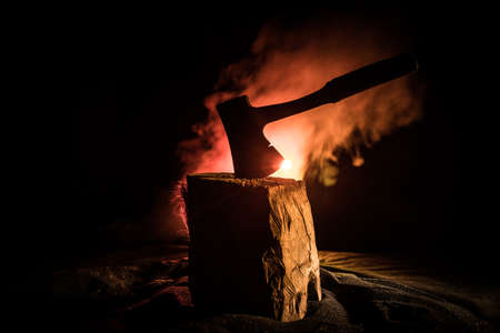 Old ax attached to the tree trunk on horror red foggy background. Scary Halloween theme with maniac...