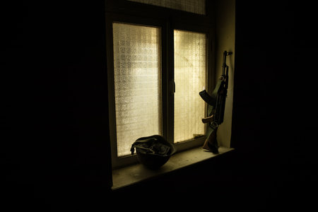 Conceptual photo of war between Russia and Ukraine. Russian weapon and helmet on windowsill at night. Old creepy room with window. Explosion outside.の写真素材
