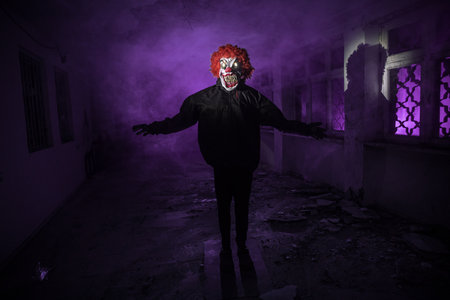 Scary evil clown with red hair standing inside ruined hall in abandoned building at night. selective focusの写真素材