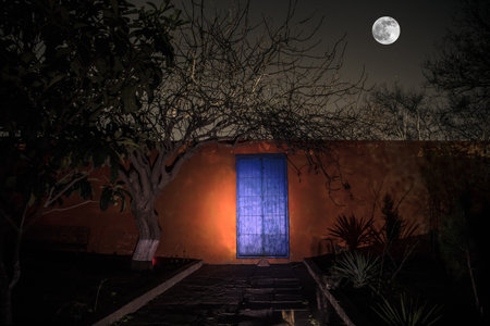 Full moon over quiet village at night. Orange wall with blue metallic Door at night. Haunted horror atmosphere. long exposure shotの写真素材