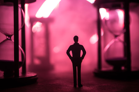 Time concept. Silhouette of a man standing between hourglasses with smoke and lights on a dark background. Surreal decorated pictureの写真素材