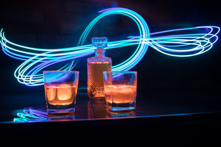Whiskey drink concept. Glass of whiskey and ice with color light and fog on dark bar background. selective focusの写真素材