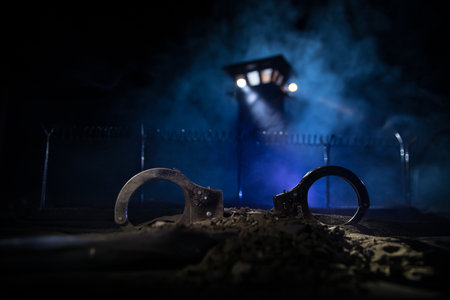 criminal justice concept. Handcuffs and Old prison watchtower protected by wire of prison fence at night. creative art decoration. selective focusの写真素材