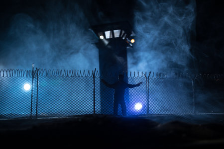 Criminal justice concept or prison break concept. Silhoette of prisoner standing near prison fence with watchtower at night. creative art decoration. selective focusの写真素材