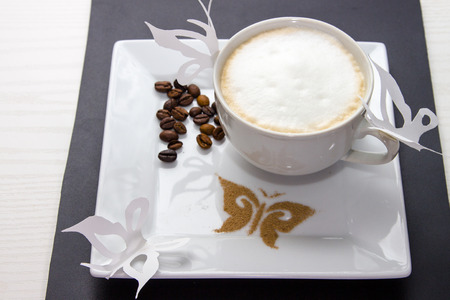 white cup of hot coffee with milk foam, cinnamon and coffee beans on the white plateの写真素材