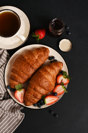 Fresh croissants with strawberries and blueberries and a cup of coffee on a dark backgroundの写真素材
