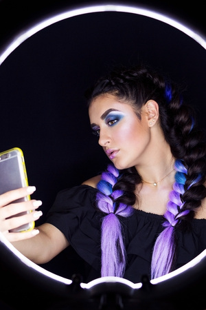 The girl makes selfie on the phone in the photo studio near the ring lamp. Studio ring light. Lamp...