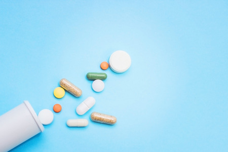 Multi-colored tablets and capsules, white bottle for tablets, pharmaceutical medicine pills on blue...
