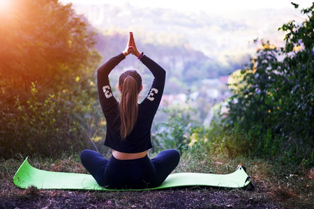 Beautiful blonde caucasian girl in green swimsuit doing asana balancing on her shoulders with twisted legs in the air on a sunny summer day in park. Yoga concept. Healthy lifestyle concept.の写真素材