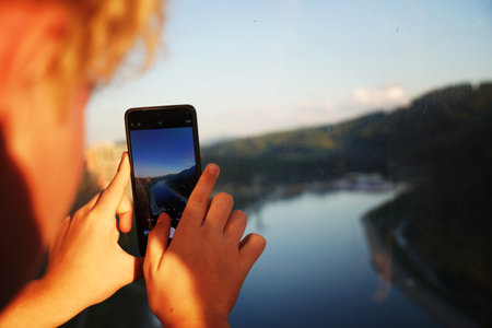 Unknown young man in red jacket takes photos with smartphone on the Lake Garda shoreの写真素材