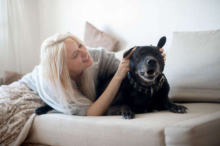 Young blonde woman and cute black dog on couch at homeの写真素材