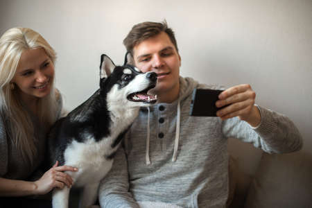 Happy young couple taking a selfie together with dog on the couch at homeの写真素材