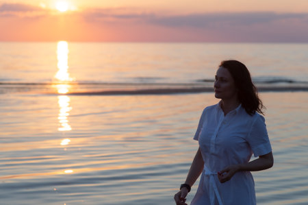 Pretty mature woman stands on the beach and looks thoughtfully into the distance at seaの写真素材