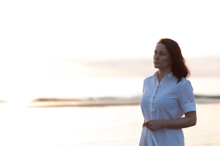 Pretty mature woman stands on the beach and looks thoughtfully into the distance at seaの写真素材