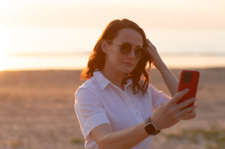 Young woman taking selfie through smart phone while standing at beach during sunsetの写真素材