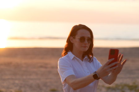 Young woman taking selfie through smart phone while standing at beach during sunsetの写真素材