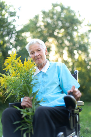 happy old senior woman sitting in a wheelchair and holding a bouquet of yellow wildflowersの写真素材
