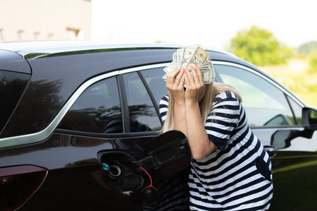 Desperate woman hides face behind cash near an open car fuel tank, concept of rising fuel pricesの写真素材