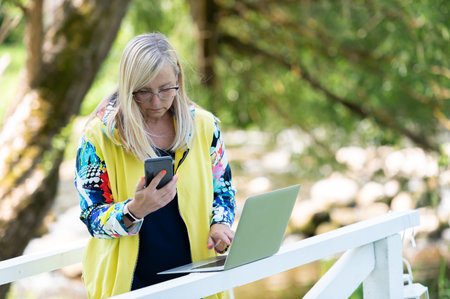 Portrait of busy sales woman standing at business park and using her laptop while writing text on mobile. Businesswoman working online.の写真素材