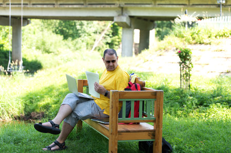 Middle-aged man man sitting on the wooden bench in park and using laptop, working with documents outdoorsの写真素材