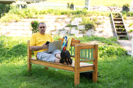 Image of content european man 50s in casual clothing sitting on bench in park and talking on cell phone while using silver laptopの写真素材