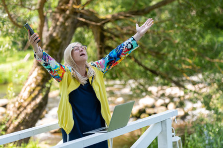 Desperate angry business woman shouting in anger with hands raised in the air, using phone and laptop outdoorsの写真素材