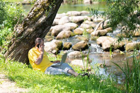 Middle aged freelancer using a laptop computer and phone while working in beautiful nature under the tree on the bank of the riverの写真素材