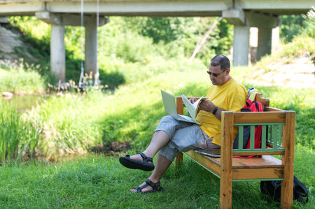 Middle-aged man man sitting on the wooden bench in park and using laptop, working with documents outdoorsの写真素材