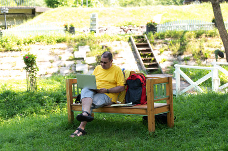 Middle-aged man man sitting on the wooden bench in park and using laptop, working outdoorsの写真素材