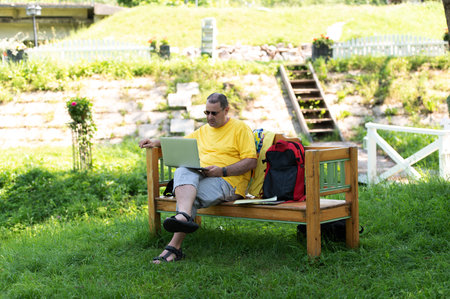 Middle-aged man man sitting on the wooden bench in park and using laptop, working outdoorsの写真素材