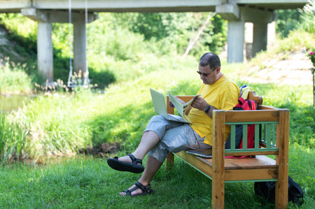 Middle-aged man man sitting on the wooden bench in park and using laptop, working with documents outdoorsの写真素材