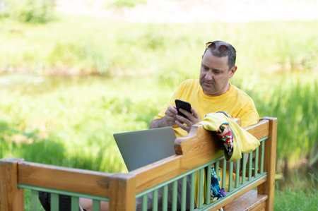 Image of content european man 50s in casual clothing sitting on bench in park and talking on cell phone while using silver laptopの写真素材