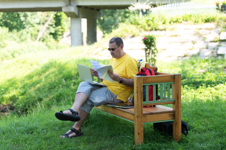 Middle-aged man man sitting on the wooden bench in park and using laptop, working with documents outdoorsの写真素材