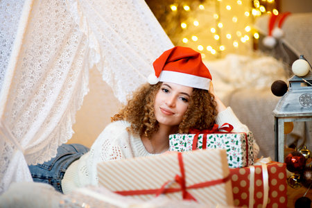 Pretty lady woman laying on Christmas giftboxes  wear knitted pullover and santa hat on bokeh lights background in decorated home roomの写真素材