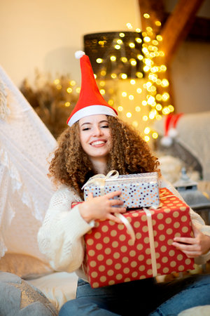 Pretty lady woman holding large Christmas giftboxes in arms wear knitted pullover and santa hat on bokeh lights backgroundの写真素材