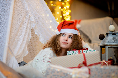 Pretty lady woman laying on Christmas giftboxes  wear knitted pullover and santa hat on bokeh lights background in decorated home roomの写真素材