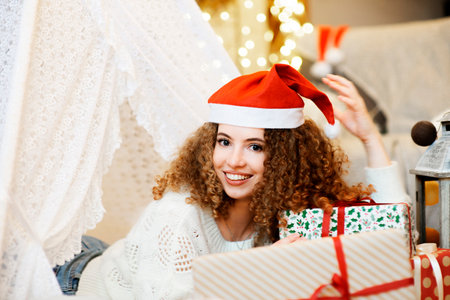 Pretty lady woman laying on Christmas giftboxes  wear knitted pullover and santa hat on bokeh lights background in decorated home roomの写真素材