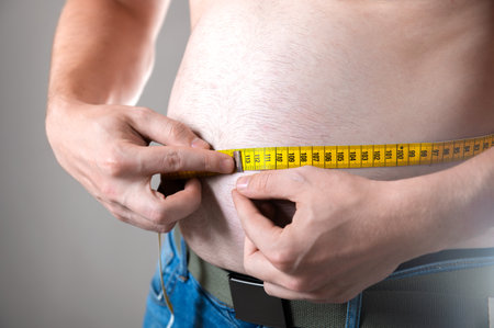 A man measures his fat belly with a measuring tape on a gray backgroundの写真素材