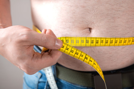 A man measures his fat belly with a measuring tape on a gray backgroundの写真素材
