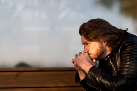 Pensive serious young bearded man wear black leather jacket sitting and thinking outdoorsの写真素材