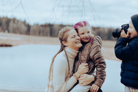 little boy with camera takes pictures of his mother and sister in nature by the lake