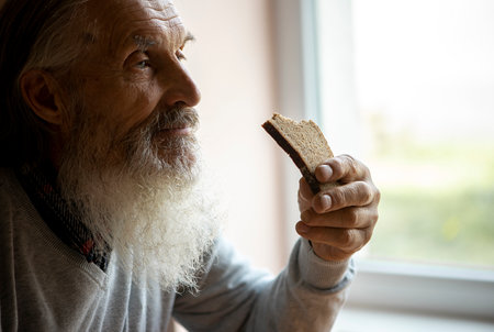Old sad man with a long gray beard sitting by the window and eating breadの写真素材
