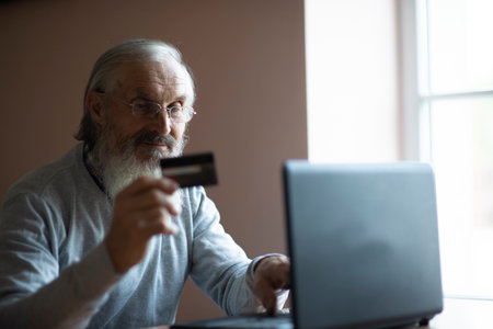 Portrait of bearded senior elderly retired man sitting at the  laptop computer and  holding credit bank cardの写真素材