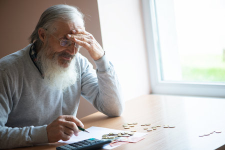 Old bearded senior man with calculator and bills counting euro money and writing notes on white sheet of paper.の写真素材